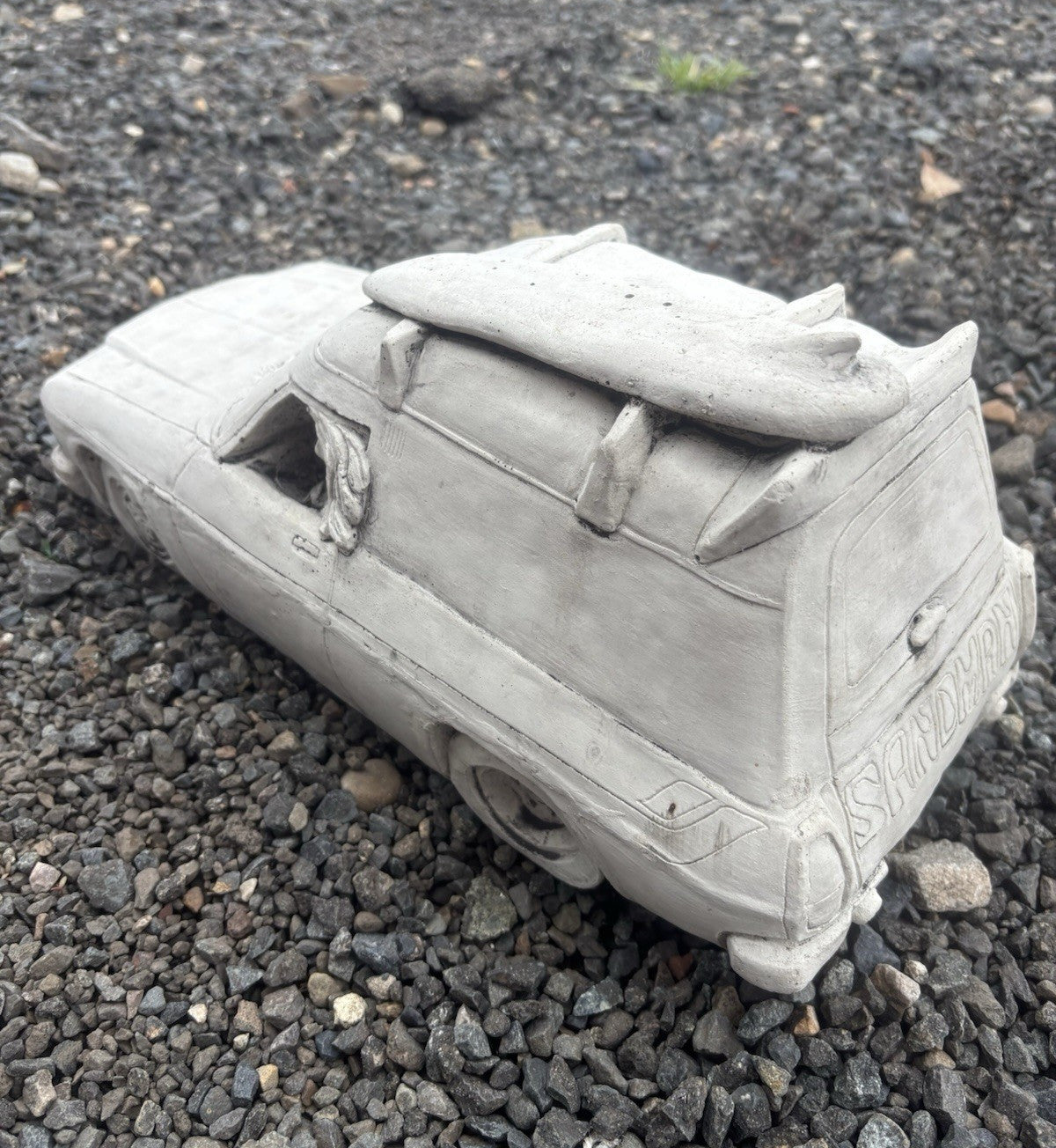 22nice4me Garden Statues concrete Sandman panel van ornament with roof surfboard on a gravel surface