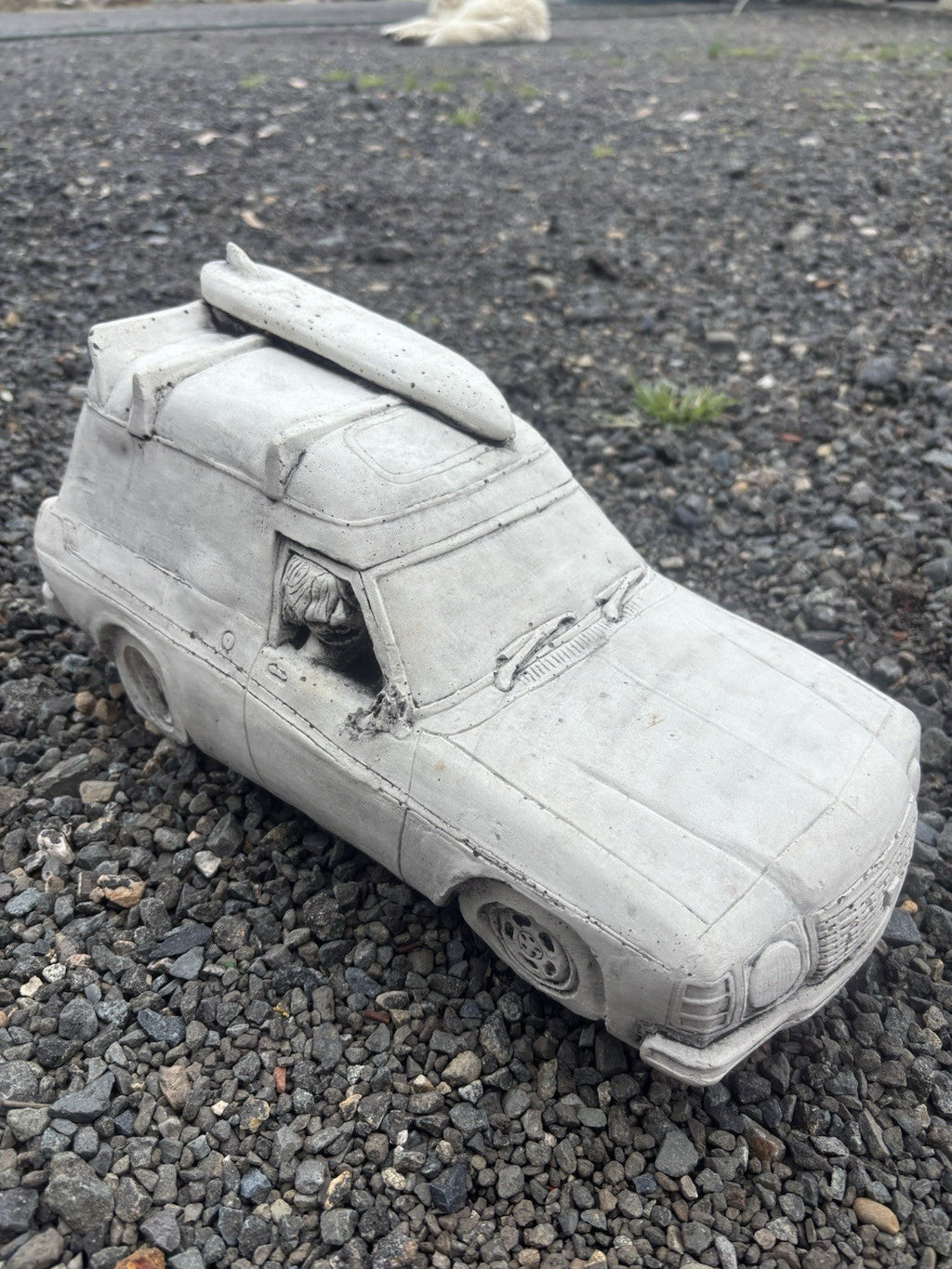 22nice4me Garden Statues concrete Sandman panel van ornament with a roof-mounted surfboard on a gravel surface
