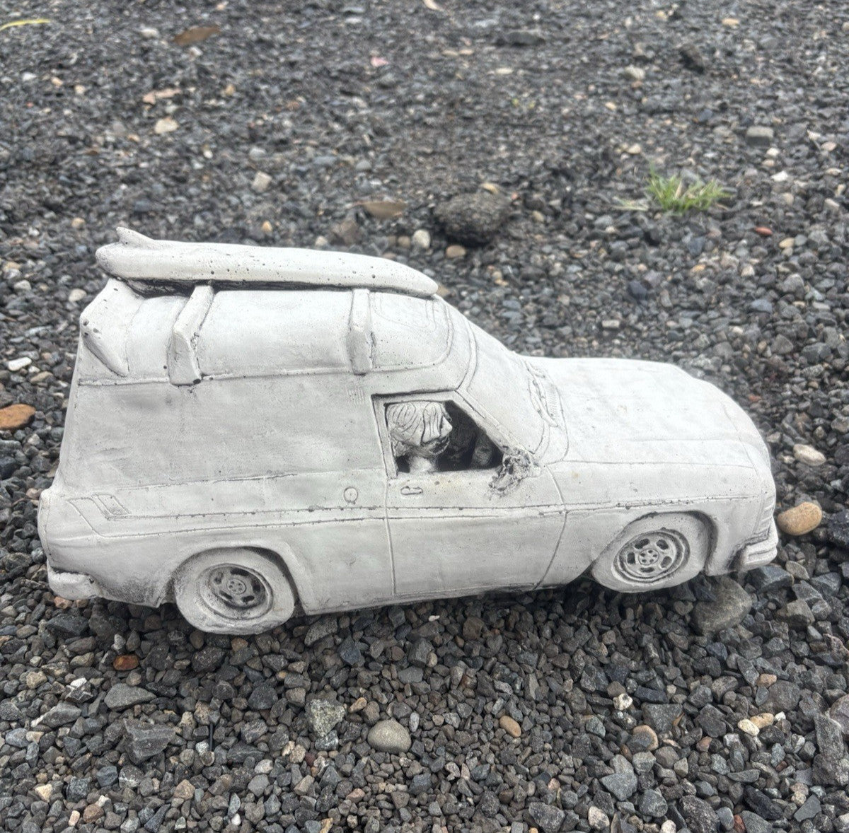 22nice4me Garden Statues concrete Sandman panel van ornament with a roof-mounted surfboard on a gravel surface