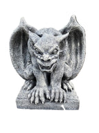 A gray stone-gothic gargoyle garden statue with wings spread, positioned outdoors on a bed of mulch with green foliage in the background.